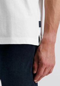 Weißes Baumwoll-T-Shirt mit seitlichen Schlitzen und einem marineblauen Etikett am Saum, kombiniert mit dunkelblauen Jeans, die einen lässigen Stil präsentieren.