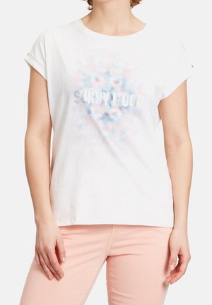 Femme portant un T-shirt blanc aux manches retroussées et un motif floral pastel avec le texte "SUNNY MOOD", assorti à un pantalon rose clair.