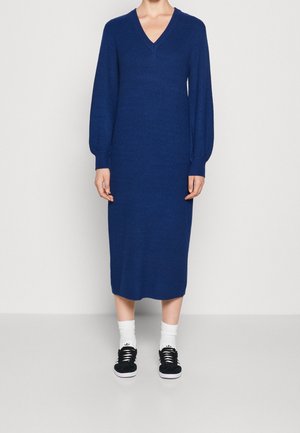 Femme portant une robe longue à manches longues, côtelée bleu jusqu'au genou avec col en V, chaussettes blanches et baskets noires, debout devant un fond uni.