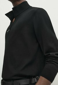 Pull noir zippé avec un col montant, fabriqué dans un tissu lisse ; doté de manches longues et d'une coupe ajustée.