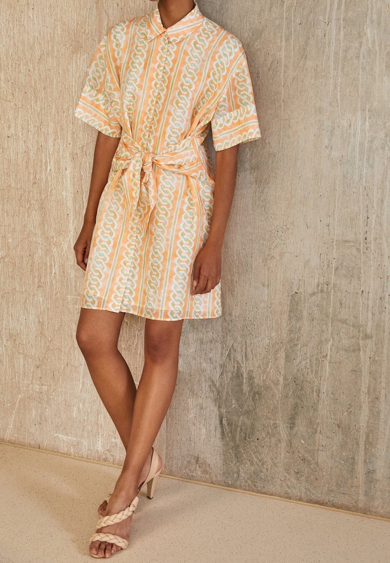 Košilové šaty s krátkými rukávy v pastelové oranžové barvě s řetízkovým vzorem, stažené v pase. Kombinované s béžovými plétanými sandály na vysokém podpatku.