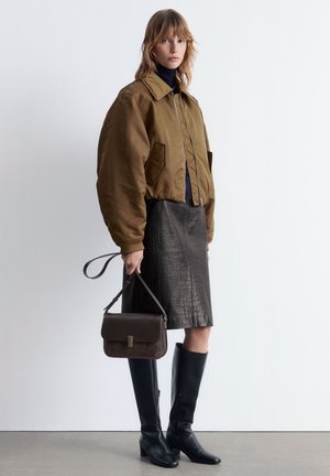 Frau trägt eine braune, cropped Jacke, einen schwarzen, strukturierten Lederrock, schwarze kniehohe Stiefel und hält eine braune Handtasche mit Tragegurt an ihrer Seite.