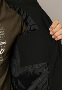 Veste noire avec un intérieur lisse et brillant et une poche intérieure ; portée sur une chemise verte olive avec un texte graphique blanc.