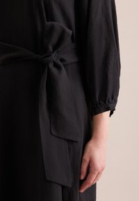 Dettaglio di un vestito di lino nero con una cintura annodata in vita. Il tessuto ha una consistenza morbida e le maniche sono gatherate ai polsini.