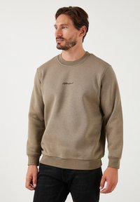 Sudadera verde oliva de tela suave, con cuello redondo y mangas largas. Detalle del logo negro en el pecho. Textura suave en toda la prenda.