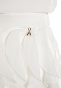 Tejido de chifón blanco en capas con bordes en forma de concha y un pequeño charm en forma de insecto dorado sujeto cerca del borde superior.