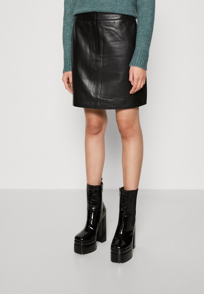 Femme portant une mini-jupe en cuir noir et des bottines noires brillantes à semelle compensée avec talons épais.