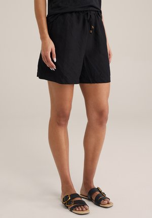 Shorts en lin noirs avec taille élastique et cordon de serrage, dotés de poches latérales. Associés à des sandales noires avec des boucles dorées.