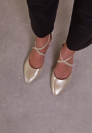 Gold, metallic ballet flats with a pointed toe and crisscross strap design. Smooth texture with subtle shine, paired with black trousers.