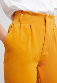 Högmidjade byxor i ljusgul tyg med veck och sidofickor, med en slät textur och en strukturerad design.