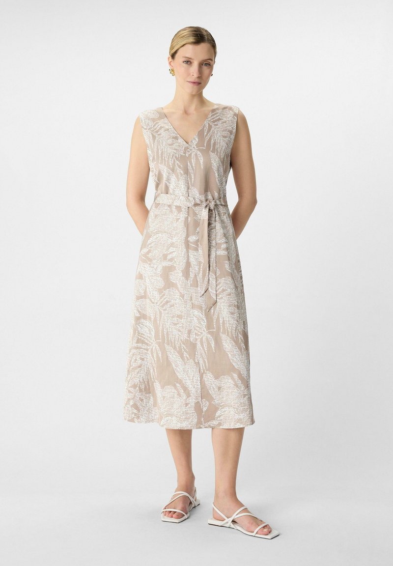 Jasno beżowa sukienka z białym wzorem kwiatowym, dekolt w serek, bez rękawów, z wiązaniem w talii i długości midi; zestawiona z białymi sandałami na paski.