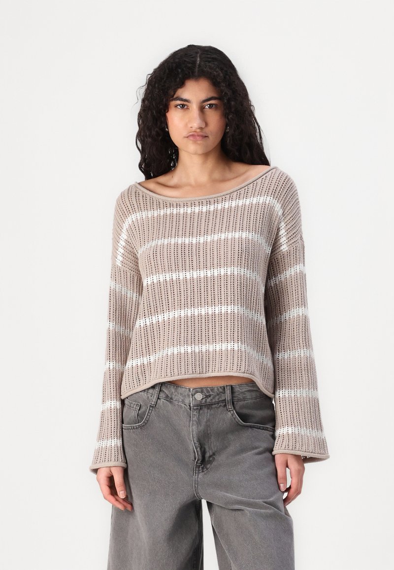 Beige stickad tröja med vid halsringning och långa ärmar, försedd med horisontella vita ränder och en lös passform, kombinerad med grå jeans.