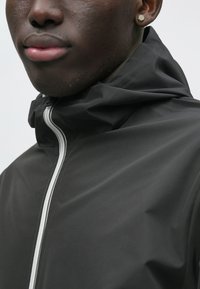 Μαύρο μπουφάν με κουκούλα από λείο ύφασμα, με λευκό φερμουάρ στο κέντρο και χαλαρή σχεδίαση στον γιακά.