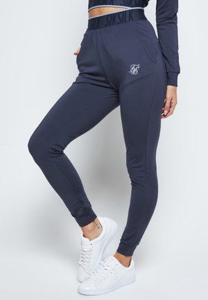 Femme portant un jogger slim bleu foncé avec taille élastique et logo, associé à des baskets blanches, debout sur un fond uni.