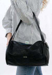 Schwarze Lederhandtasche mit strukturiertem Oberflächen, ausgestattet mit zwei langen Trägern und einem silbernen Logo. Getragen über einem gestreiften Hemd und Jeans.