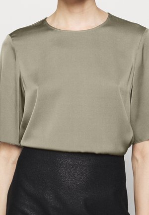 Blouse en satin à manches courtes de couleur olive douce. Encolure ronde et texture douce avec une coupe ample. Associée à une jupe noire texturée.