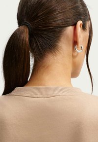 Μπεζ πουλόβερ με ριμπ λαιμόκοψη, φορεμένο με ασημένιο κρίκο σκουλαρίκι, τα μαλλιά είναι χτενισμένα σε χαμηλή αλογοουρά, παρουσιάζοντας φυσική υφή μαλλιών.