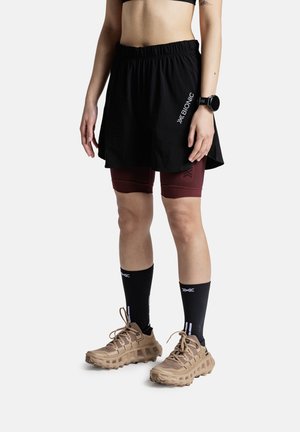 Personne portant une jupe de sport noire avec un short de compression bordeaux, des chaussettes noires, des chaussures de trail beiges et une montre connectée noire au poignet.