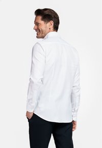 Chemise blanche à manches longues avec col pointu, poignets boutonnés et ourlet arrondi. Le tissu semble lisse et léger.