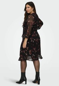 Mujer llevando un vestido negro de longitud media con patrones florales en burdeos y blanco, mangas transparentes, medias negras y botines de tacón negros, de pie en actitud de perfil.