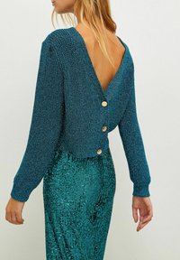 Woman wearing a teal knit sweater with gold buttons down the open back paired with teal sequin pants, standing against a plain background.