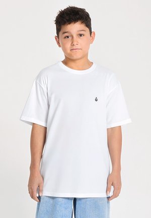 Garçon aux cheveux courts et foncés, portant un t-shirt blanc uni et un short en jean bleu clair, debout devant un fond blanc.