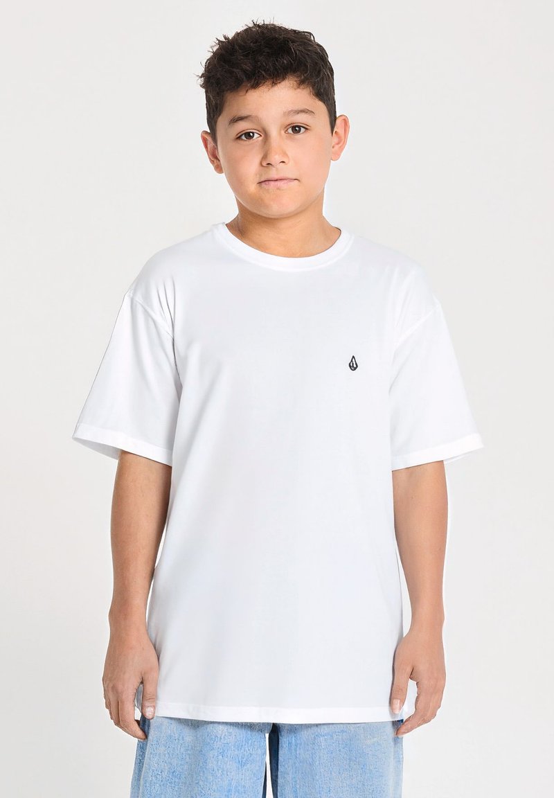 Garçon aux cheveux courts et foncés, portant un t-shirt blanc uni et un short en jean bleu clair, debout devant un fond blanc.