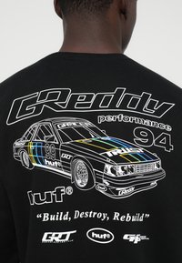 Schwarzes T-Shirt mit weißer Linienzeichnung eines Rennwagens, nummeriert 99, und Text: "GReddy Performance 94," "Bauen, Zerstören, Wiederaufbauen," sowie Markenlogos.