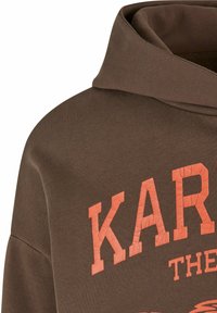 Sudadera de algodón marrón con un gran estampado naranja agrietado que dice "KAR." Cuenta con una capucha amplia y un tejido texturizado.