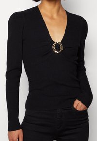 Schwarzes, geripptes Langarmshirt mit tiefem V-Ausschnitt, das ein goldenes, rundes Detail an der Brust und eine figurbetonte Silhouette aufweist.