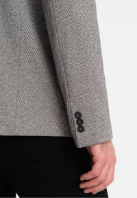 Blazer a spina di pesce grigia con vestibilità su misura, rever a punta e tre bottoni neri ai polsini, abbinato a una camicia nera.