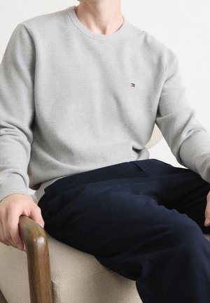 Grau strukturierter Sweatshirt mit Rundhalsausschnitt und dezentem Logo. Kombiniert mit dunkelblauen Hosen. Die Person sitzt und hält sich am Armlehne eines Holzstuhls fest.