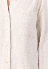 Light beige linen shirt with a button-up front, single chest pocket, and a textured fabric. Features rounded collar and long sleeves.