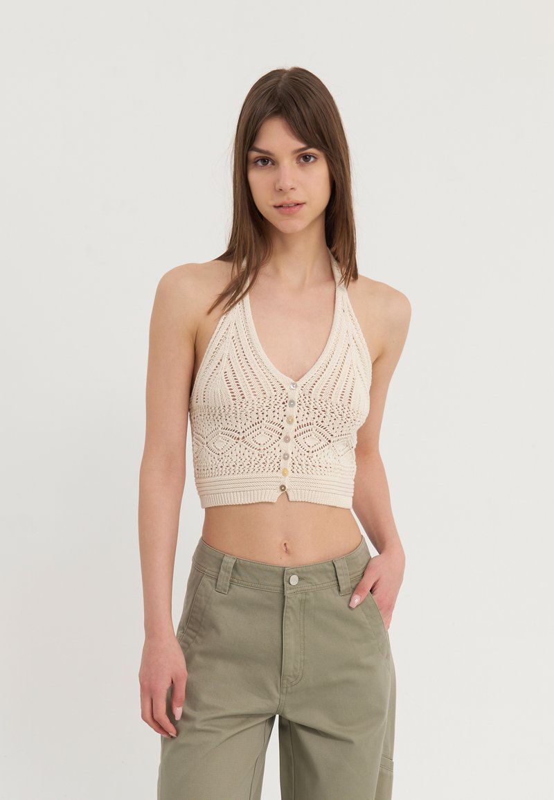 Kvačkasti top na halter u off-white boji, s V-izrezom, zatvaranjem na dugme i složenim uzorcima. Uparen s hlačama visokog struka u zelenu boju.