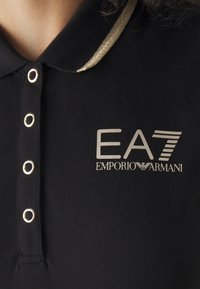 Svart polo shirt med en glittrande guldkrage, prydd med silverfärgade EA7- och Emporio Armani-loggor. Metallknappar framhäver knapparna.