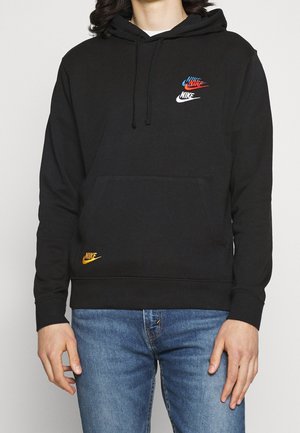 Persoon draagt een zwarte Nike-hoodie met blauwe, rode, witte en gele Nike-logo's, gecombineerd met een blauwe spijkerbroek, staand tegen een effen achtergrond.