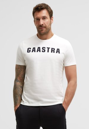 Man wearing a white short-sleeve T-shirt with "GAASTRA" text, black pants, and a tattoo sleeve on his left arm, hands in pockets.