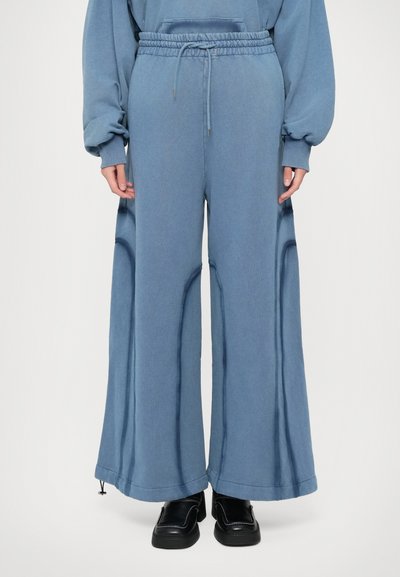 Personne portant un pantalon large bleu avec une taille froncée et un cordon de serrage, associé à des chaussures noires à enfiler avec des coutures blanches.