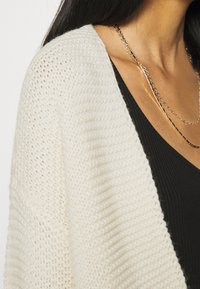 Knitted white cardigan with a loose fit, featuring a textured pattern and open front design, layered over a black ribbed top.