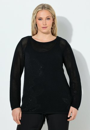 Kobieta o blond włosach, ubrana w czarny, dziergany sweter z długim rękawem z subtelnym kwiatowym wzorem oraz czarne spodnie, stojąca na tle białej ściany.