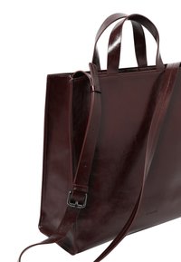 Sac tote en cuir brun foncé brillant avec deux poignées et une bandoulière réglable, vue latérale angulaire sur fond blanc.