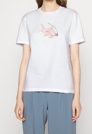 Kobieta nosząca biały T-shirt z nadrukiem na piersi różowej ryby wydmuchującej bąbelki i mającej na sobie maskę do nurkowania, w połączeniu z niebieskimi spodniami.