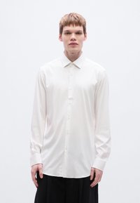 Λευκό πουκάμισο με μακριά μανίκια και γιακά με κουμπιά, λείο ύφασμα και άνετη γραμμή· διαθέτει κλασικό κλείσιμο με κουμπιά και μανσέτες με ένα κουμπί.