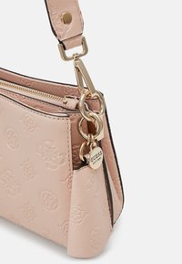 Sac à main rose clair avec logos embossés, quincaillerie dorée, fermeture zippée, et un charm rond de la marque Guess suspendu à l'attache de la sangle.