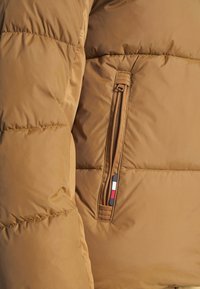 Brown puffer jacket with quilted design, featuring a zippered pocket on the left sleeve and a multi-colored branding accent.