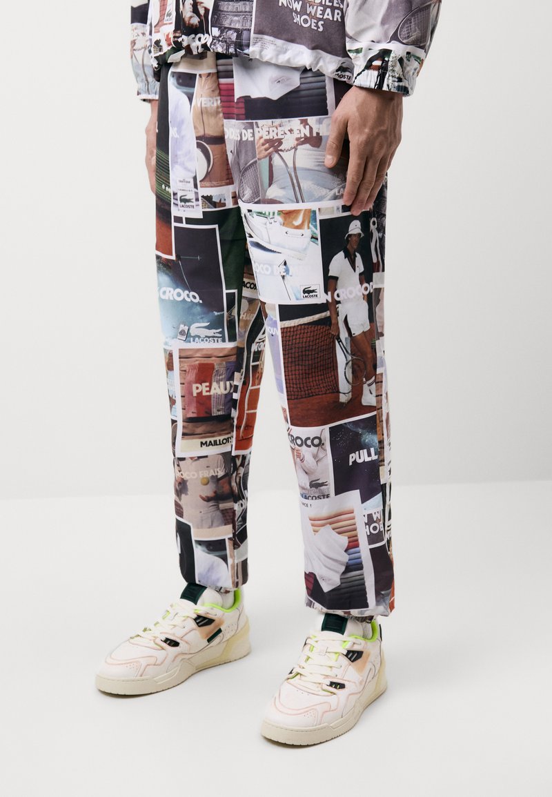 Hombre que lleva pantalones con un estampado de collage fotográfico colorido con temática de tenis y zapatillas de color claro con acentos en verde neón, de pie sobre un suelo blanco.