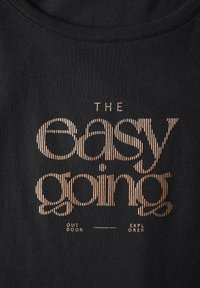 Černé bavlněné tričko s zlatým textem "THE easy going" s "OUT DOOR" a "EXPLORER" pod ním, na texturovaném pozadí.