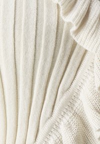 Krémsvětlá pletená látka s vertikálním žebrováním a volánovými detaily. Povrch vykazuje variace v pletených vzorech, což dodává hloubku.