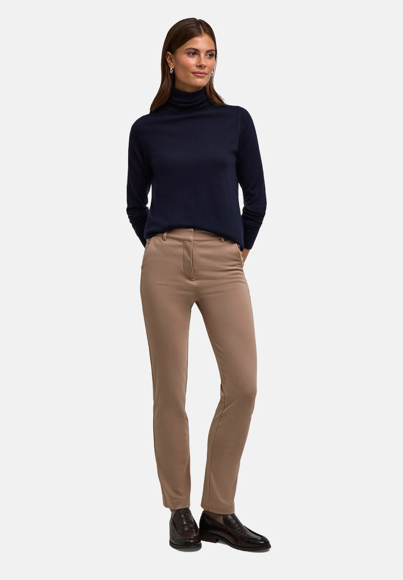 Marineblauer Rollkragenpullover kombiniert mit hellbraunen Hose; die Hose hat eine taillierte Passform mit Seitentaschen und wird durch dunkle Dresserschuhe ergänzt.