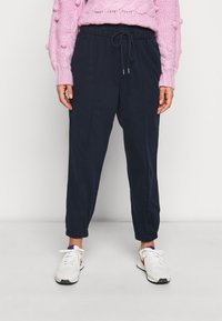 Námorně modré joggingové kalhoty s elastickými manžetami, stahovací šňůrkou v pase a postranními švy. Stylizováno s růžovým pleteným svetrem a bílými teniskami.